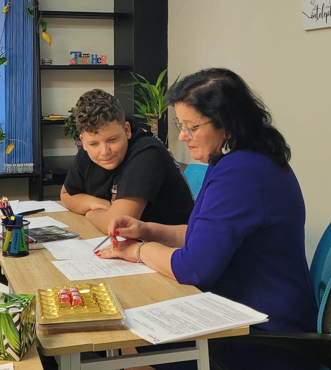 Profesoară explicând unui elev de gimnaziu, aplecați amândoi peste aceeași foaie
