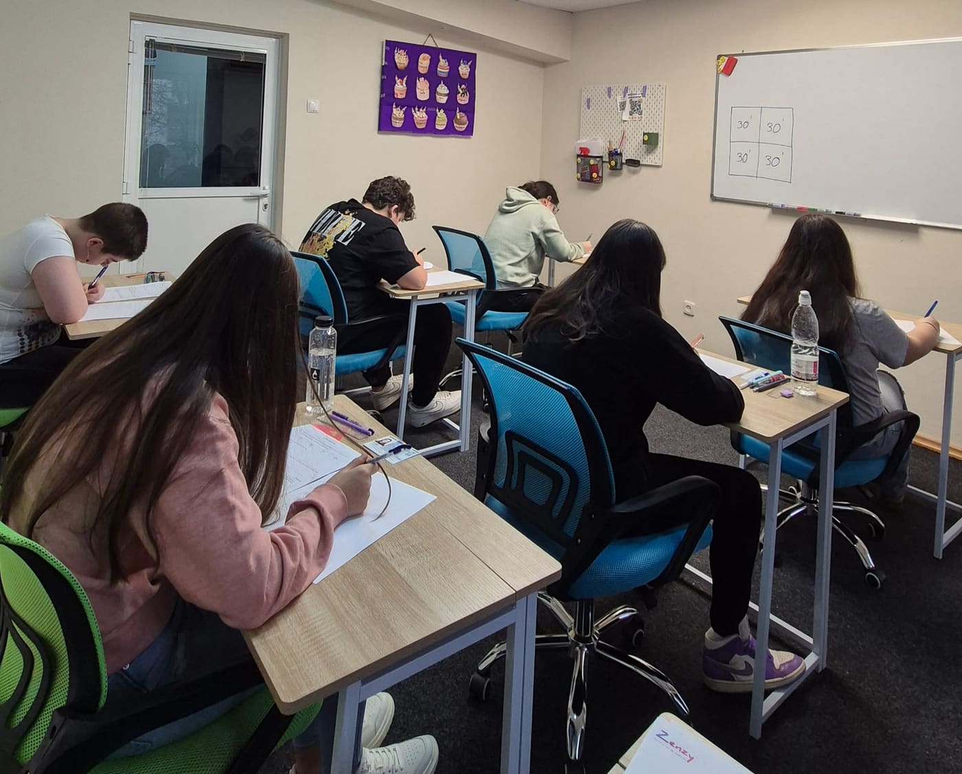 Elevi scriind la simulare, fiecare la banca lui, cu timpul pe probe notat pe tablă